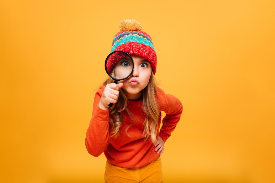 joyful-young-girl-in-sweater-and-hat-looking-at-the-camera-with-magnifier-over-orange_171337-1729.jpg